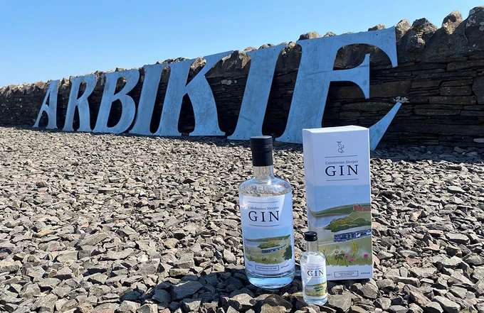 Two bottles and a box of gin displayed on rocky ground with large metal letters spelling 'ARBIKIE' against a stone wall under clear blue sky.