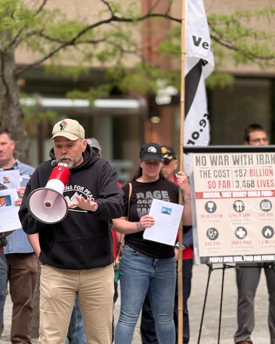 A person is speaking into a megaphone during a protest. They are wearing a cap and a "Veterans for Peace" hoodie. Nearby, another individual holds a sign that reads "No War With Iran," detailing costs and lives affected. A Veterans for Peace flag is visible in the background.