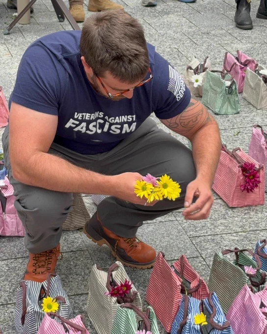 A person wearing a "Veterans Against Fascism" T-shirt is crouched down, holding yellow flowers. They are surrounded by striped bags with more flowers inside.