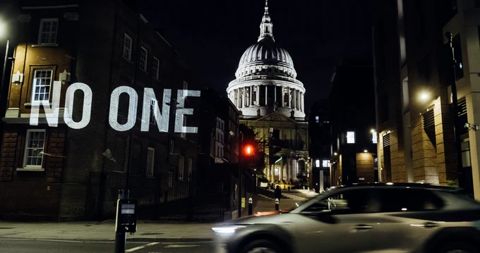 An image of the words NO ONE s projected onto a building at night with St Paul's Cathedral in the background.