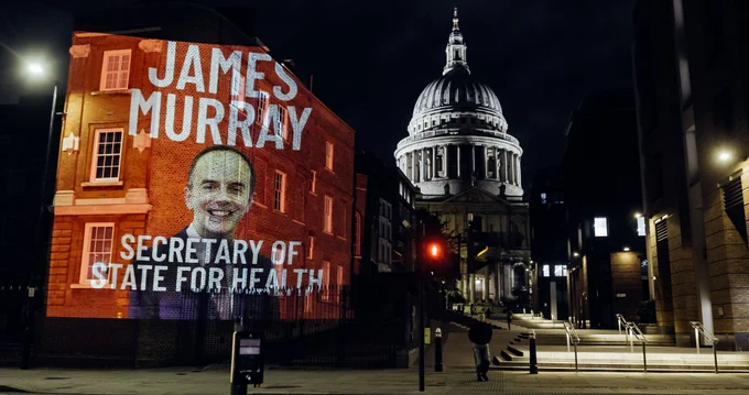 An image of James Murray with his name and Secretary of State of Health in white text on a red background is projected onto a building at night with St Paul's Cathedral in the background.