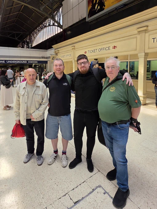 Four men pose for a photograph on the concourse of a railway station in front of the ticket office.
