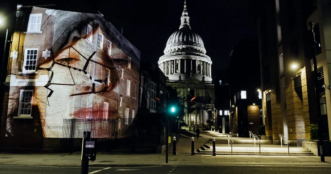 The image of a white women wearing an eye mask pushed up to her forehead is projected onto a building at night with St Paul's Cathedral in the background.