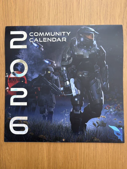 A photograph of the 2026 Halo Community Calendar by JumperScape, placed on a wooden table. The artwork on the front features Master Chief holding a pistol and walking through a dark forest on Halo with a group of Marines behind him. To the left of the cover it says, “2026 Community Calendar.”