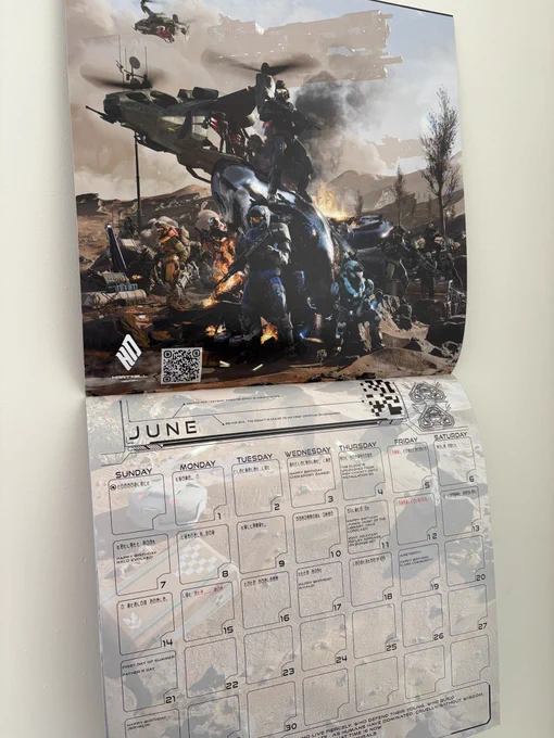 A photograph of the 2026 Halo Community Calendar, now unfurled to the June page and hung on a white wall. The artwork for June features Noble Team surrounded by battlefield wreckage on Reach.