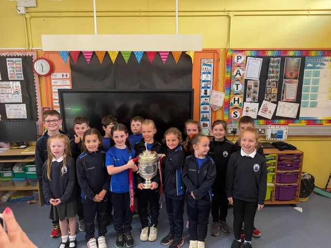 A group of children in a classroom, smiling and holding a large trophy, suggesting a celebration or achievement. The room is decorated with colorful banners and educational posters.