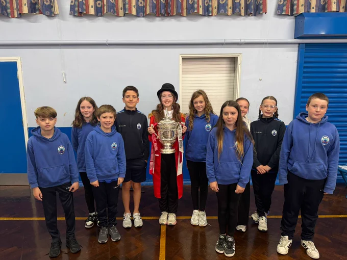 A group of students in a school gymnasium standing with a trophy. The mood is joyful and proud.