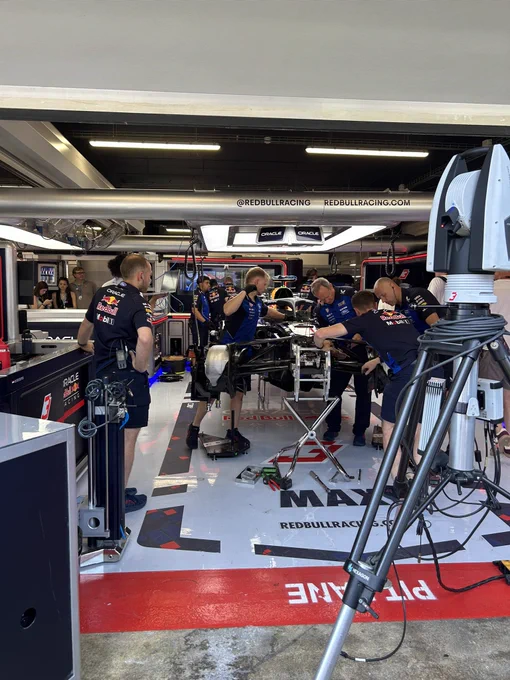 Inside the Oracle Red Bull Racing garage at the Spanish Grand Prix, mechanics in navy Red Bull and Mobil 1 team kit work on Max Verstappen’s stripped-down RB21 Formula 1 car, raised on stands with bodywork removed to expose the chassis and front suspension. A white Hexagon measuring scanner on a tripod stands in the foreground, with “PIT LANE” marked in red on the garage floor and Oracle and TAG Heuer branding overhead.