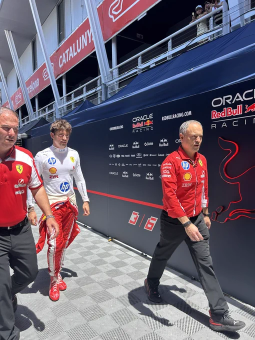 Ferrari driver Charles Leclerc walking through the paddock at the Circuit de Barcelona-Catalunya during the Spanish Grand Prix, wearing his white and red Scuderia Ferrari race suit tied down at the waist with HP, Shell, ZYN, and Puma branding. He is flanked by two Ferrari team members in red shirts, passing the navy Oracle Red Bull Racing hospitality wall covered in sponsor logos and a “3/6” graphic.