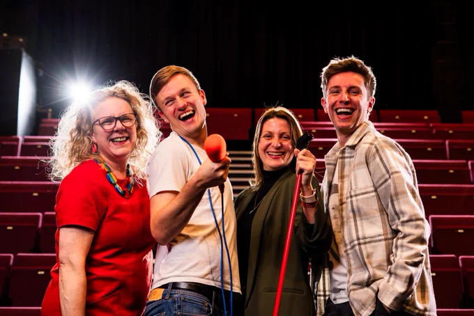 Four people are standing together in a theatre with red seats. They are smiling and appear happy. Two are holding a microphone and a microphone stand. A bright spotlight shines in the background.