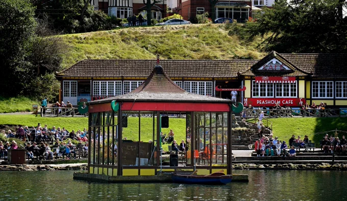 A lakeside cafe with a glass pavilion on the water, surrounded by people enjoying a sunny day in Peasholm Park Scarborough.