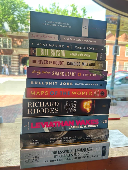 A stack of books in the window of Harvard Book Store. The books are as follows, from top to bottom:
- "The Brothers Karamazov" by Fyodor Dostoyevsky
- "And Then There Were None" by Agatha Christie
- "Anaximander" by Carlo Rovelli
- "A Walk in the Woods" by Bill Bryson
- "The River of Doubt" by Candice Millard
- "Shark Heart: A Love Storey" by Emily Habeck
- "Bullshit Jobs" by David Graeber
- "Maps of the World Quiz Book" by Hardie Grant Explore
- "The Making of the Atomic Bomb" by Richard Rhodes
- "Leviathan Wakes" by James S. A. Corey
- "True Nature: The Pilgrimage of Peter Matthiessen" by Lance Richardson
- "The Essential Peanuts by Charles M. Schulz: The Greatest Comic Strip of All Time" by Mark Evanier and Charles M. Schulz (Illustrator)