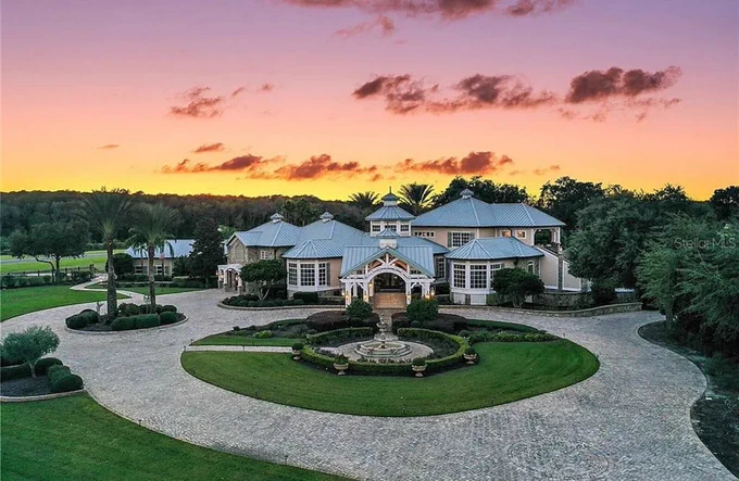 A stunning luxury mansion framed by a vibrant sunset. The circular driveway and landscaped garden enhance the grandeur of the property.
