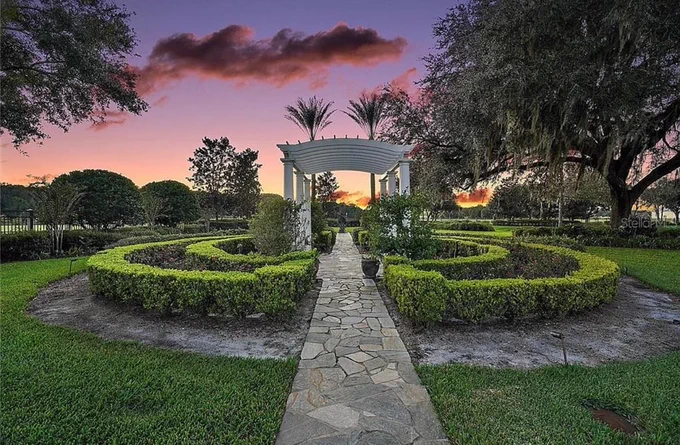 A peaceful garden with a stone path leading to a white archway surrounded by manicured hedges at sunset. The sky is a vibrant mix of purple and orange hues, adding a serene and tranquil mood.