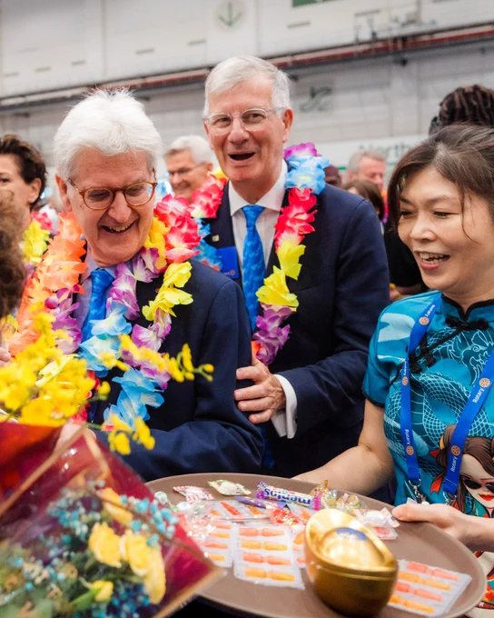 A group of people wearing colorful flower leis stand together, smiling and holding a tray with various items. They appear to be at a lively event, surrounded by a cheerful atmosphere.