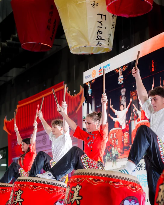 A group of performers in colorful attire energetically play large traditional drums. Behind them, a large screen displays the same performance. Hanging lanterns with Chinese characters and the word "Friendship" decorate the scene.