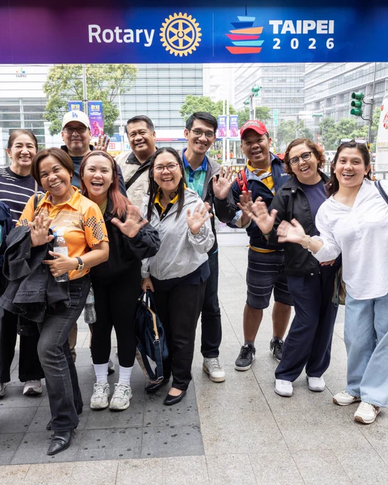 A group of people smiling and waving under a sign displaying the Rotary logo and "Taipei 2026." They are standing on a city street with buildings and trees in the background.