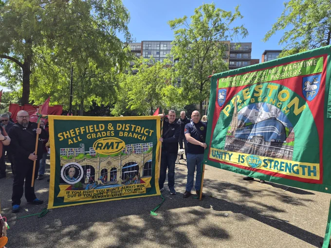 Group of people holding colorful Sheffield & District and Preston RMT union banners in a sunny park with trees and buildings in the background.