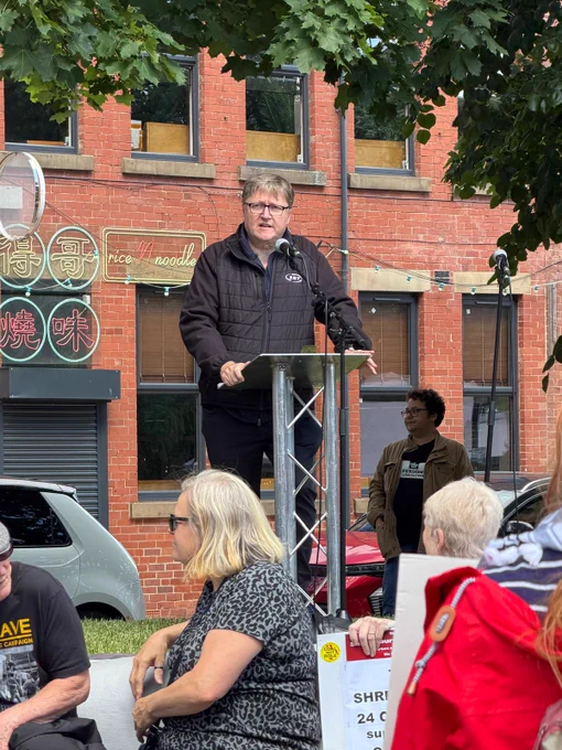 Man in black jacket speaking at outdoor podium in front of brick building with neon signs and seated audience.
