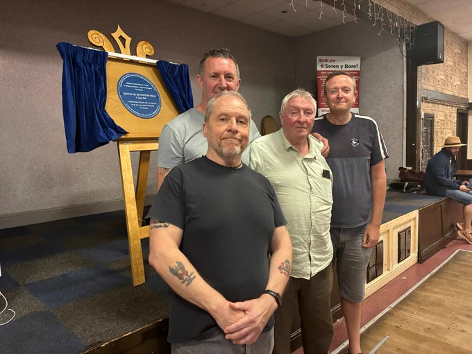 Four men standing together indoors near a large wooden plaque with blue curtains on a stand in a casual setting.