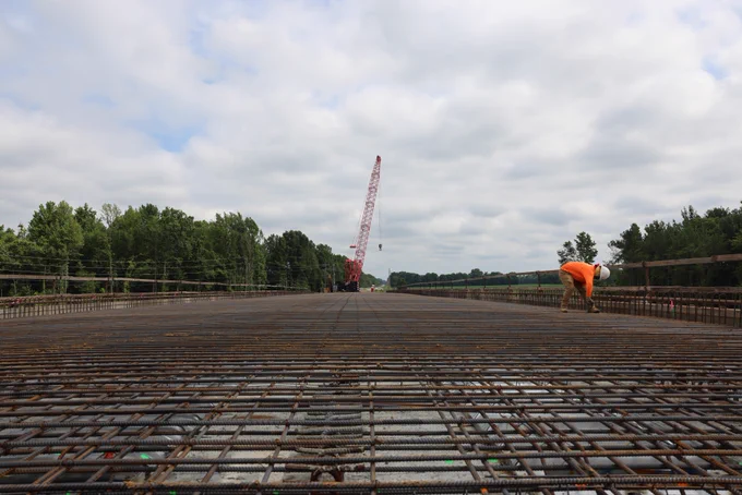 A person in an orange shirt works on a large, open expanse of rebar on a bridge construction site on Highway 51 in Madison County. In the background, there is a red construction crane amidst a landscape of trees under a partly cloudy sky.