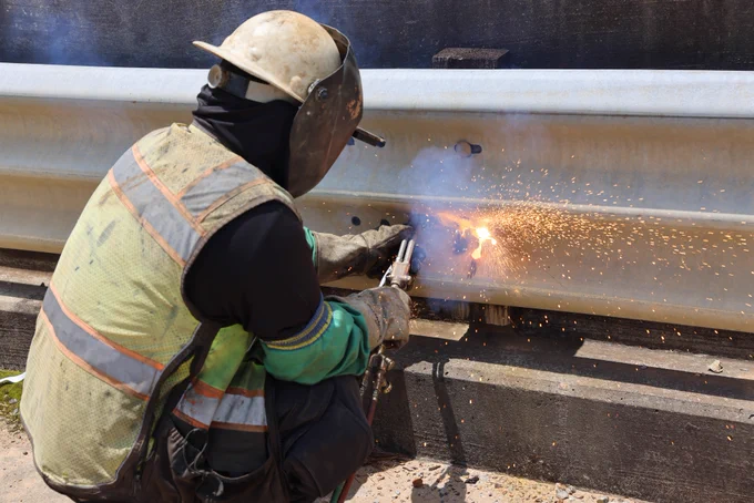 A person in protective gear is welding a metal guardrail on at a bridge replacement site along Highway 80 over the Chunky River in Lauderdale County. Bright sparks are visible.