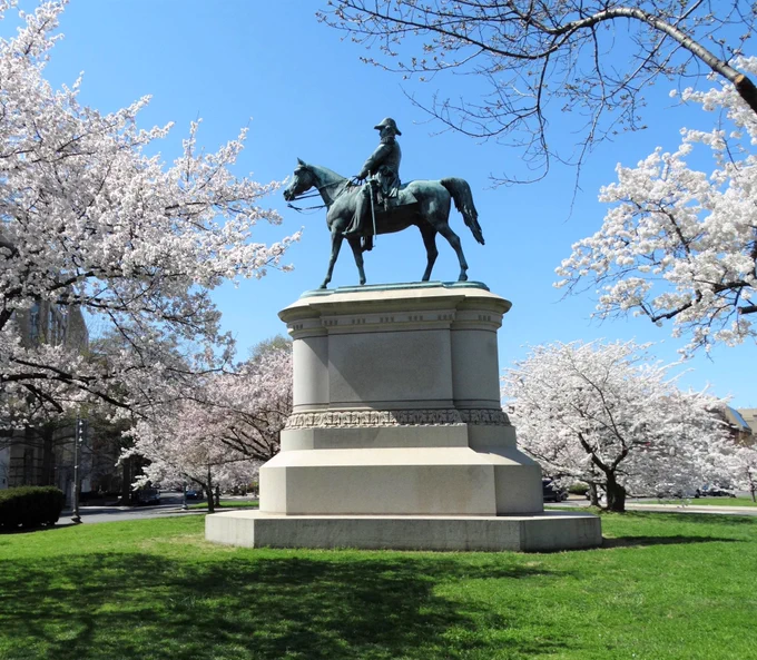 Bronze equestrian statue of a historical figure on a horse surrounded by blooming cherry blossom trees under a clear blue sky.