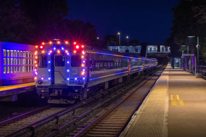 The Metro-North Knicks Lights train glowing in blue and orange as it passes by a train station in the evening.