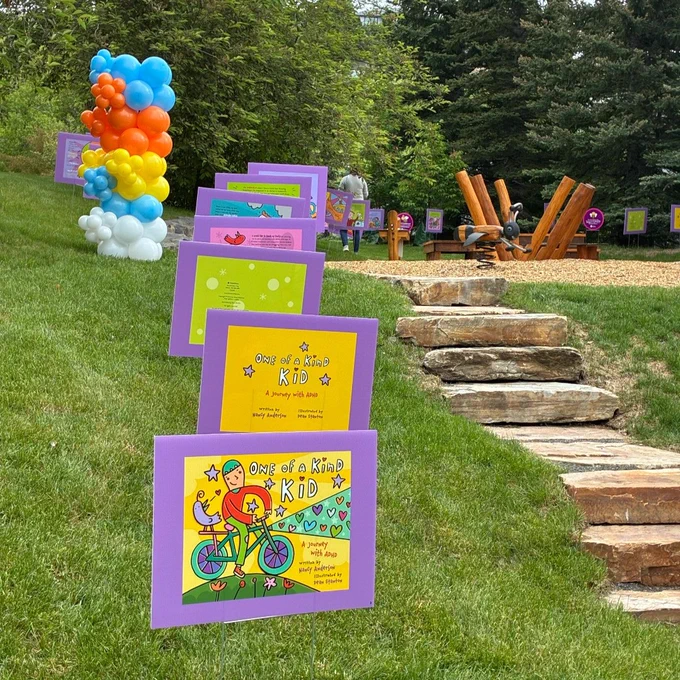 A grassy pathway features colourful, illustrated signs, leading up to a play area. Balloons in various colours are to the side, and trees are in the background.