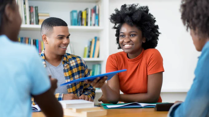 A group of students sit at a table and pass a folder to one another.