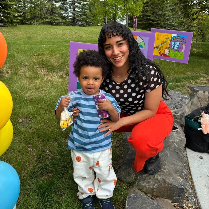 A person kneels on grass next to a child holding a snack and drink. There are colourful balloons on one side and vibrant artwork on stands in the background. Tall trees surround the scene.