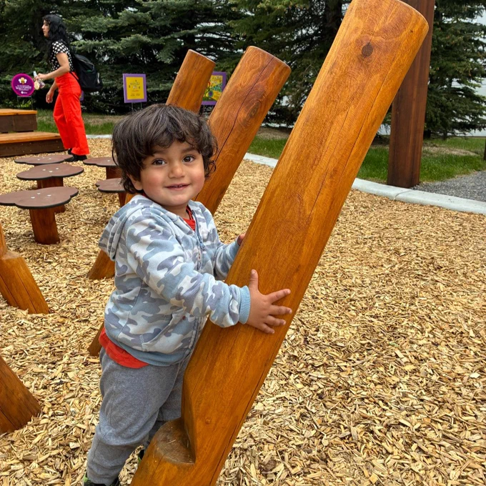 A child wearing a camouflage hoodie and gray pants is climbing on wooden play structures in a playground. The ground is covered in wood chips, and there are trees and more playground elements in the background.