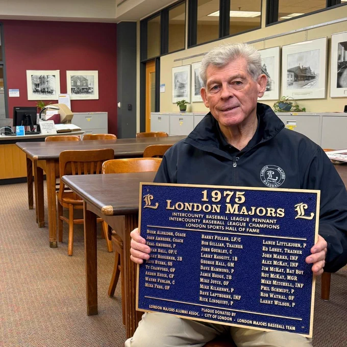 Barry Boughner sits in the London Room at Central Library holding a blue commemorative plaque recognizing the 1975 London Majors championship team. Research tables, archival storage cabinets, and historic photographs line the room behind him.