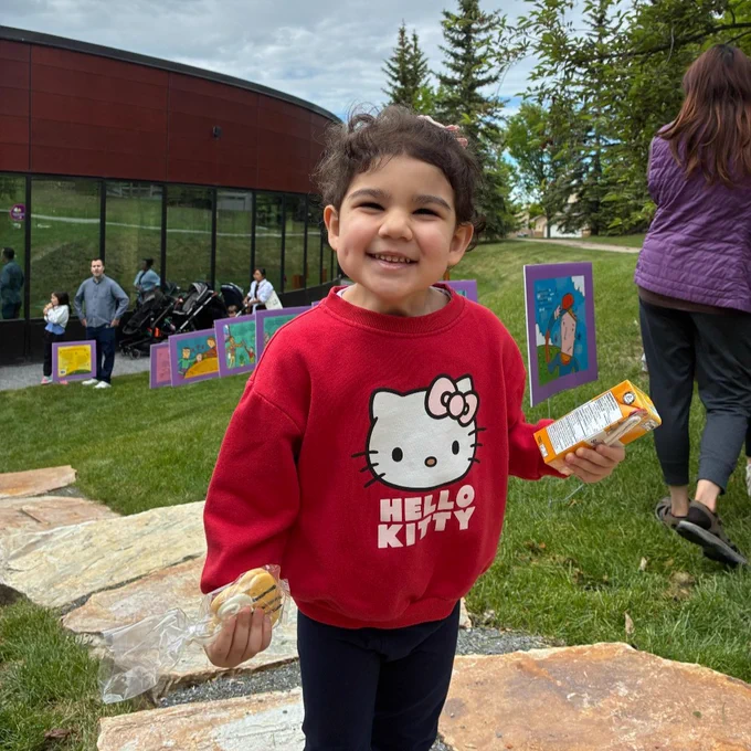 A child is standing outdoors on a grassy area, wearing a red Hello Kitty sweater and holding a snack.