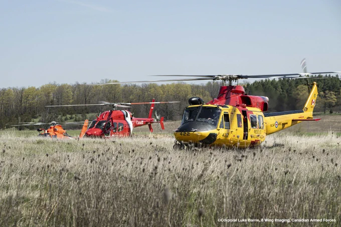 Three search-and-rescue helicopters, including one yellow and two red, parked in a grassy field with a forest in the background.