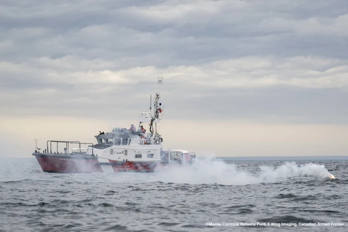 A Canadian Coast Guard vessel sails at sea while a smoke distress signal floats nearby on the water.