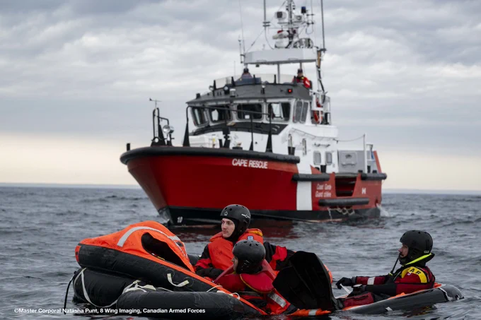 Rescue personnel in survival suits float in the water near a life raft, with a Canadian Coast Guard vessel in the background.