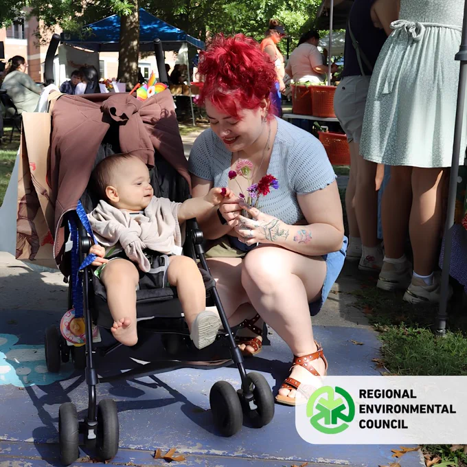 Woman with red hair kneeling beside a baby in a stroller, offering purple flowers at the REC Farmers Market at University park. 