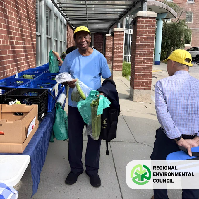 Man in a yellow cap and blue shirt holding fresh vegetables in green bags at an REC Mobile Farmers Market Location.