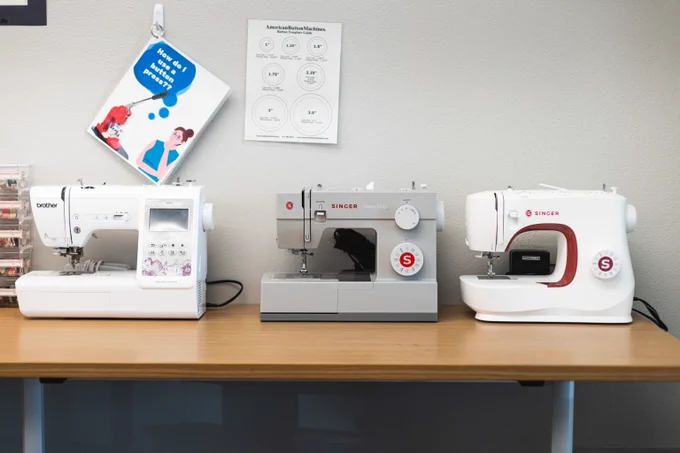 Three sewing machines from Brother and Singer brands arranged side-by-side on a wooden table against a wall in the Belle Isle Library Makerspace.