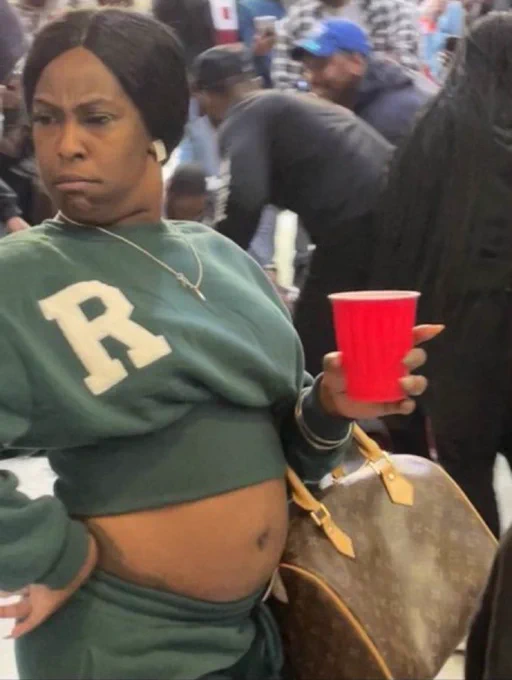 A woman in a green crop top sweat suit holding a red solo cup and LV purse. Looking flummoxed, mildly concerned, and a tiny tiny tiny bit annoyed.