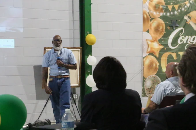Man in blue shirt and jeans speaking into a microphone at a decorated indoor event with audience seated.