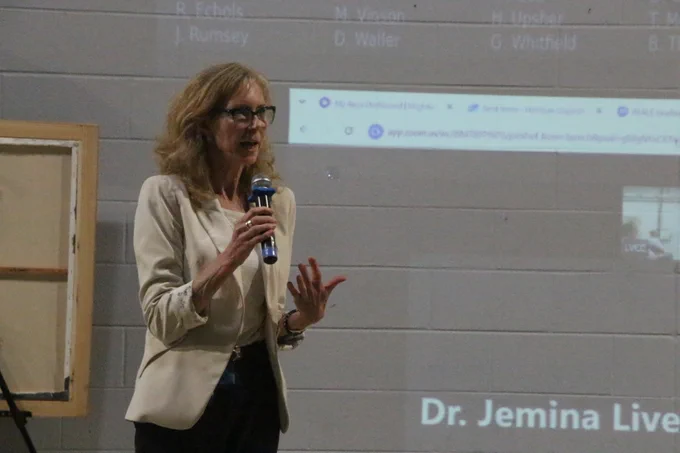 Woman in a beige blazer holding a microphone and speaking in front of a projected presentation slide on a gray wall.