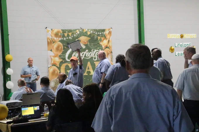 A group of people in blue shirts gathered indoors celebrating with a 'Congrats Grad' banner and gold balloons in the background.