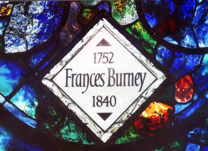 A stained glass window featuring a diamond-shaped panel with the text "1752 Frances Burney 1840" inscribed in dark lettering. The surrounding glass is vibrantly colored, with patches of blue, green, and red.