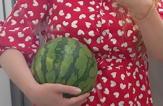 Man sieht den Oberkörper einer Frau mit einem roten Kleid und weißen Herzen drauf. Im Arm trägt sie eine ganze Wassermelone
