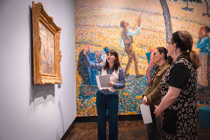 A person gestures toward a framed painting in a gallery, speaking to two other individuals. A large, colorful mural depicting people and trees covers the background wall.
