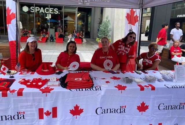 Groupe de 5 personnes portant des chemises rouges sur le thème du Canada, assises et debout derrière une table couverte de drapeaux canadiens et de marchandises sous une tente.