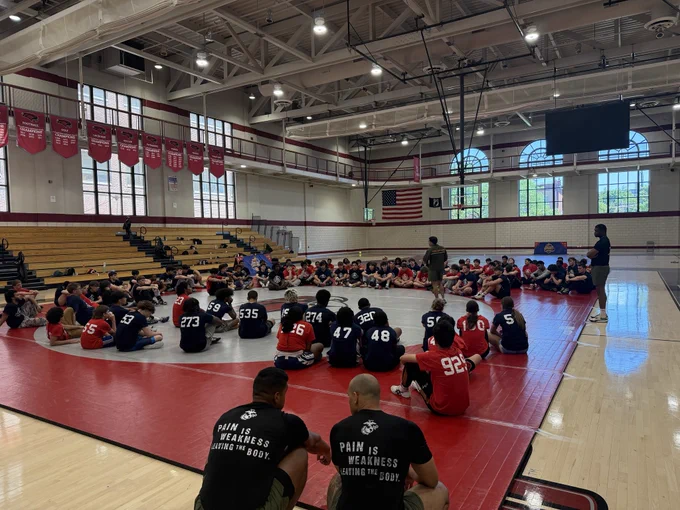 A group of people, wearing numbered jerseys, sit in a circle in a gym. Two instructors are leading the session, with motivational text visible on a participant's shirt.