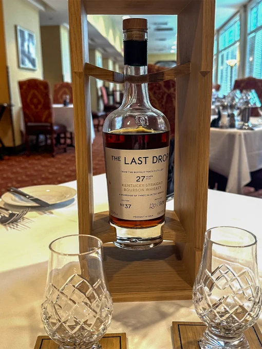 A bottle of Release No. 37: The Last Drop 27 Year Old Kentucky Straight Bourbon from Buffalo Trace Distillery sitting on the table at 1875: The Steakhouse. Two empty, intricately designed whiskey glasses are placed in front on wooden coasters. The background shows a fine dining setting with tables and chairs.